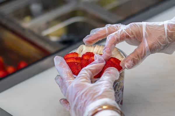 Gloved hands carefully placing sliced strawberries over banana in an acai bowl on a white prep surface.