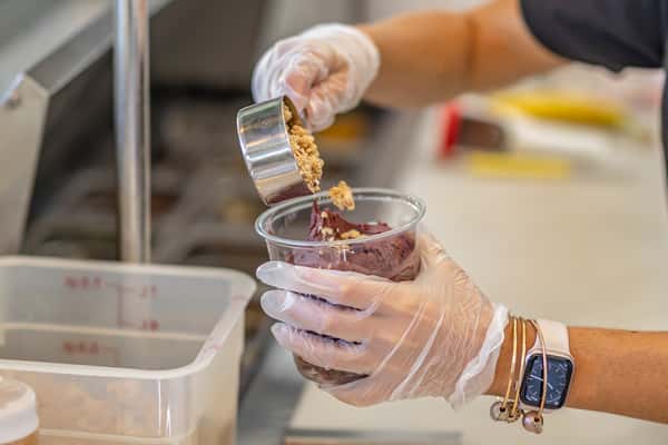 A gloved worker is adding granola from a metal measuring cup onto a freshly scooped açaí bowl. The background shows a prep station with ingredient containers.