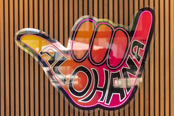 Colorful ALOHANA shaka hand sign logo mounted on a vertical wooden slat wall.