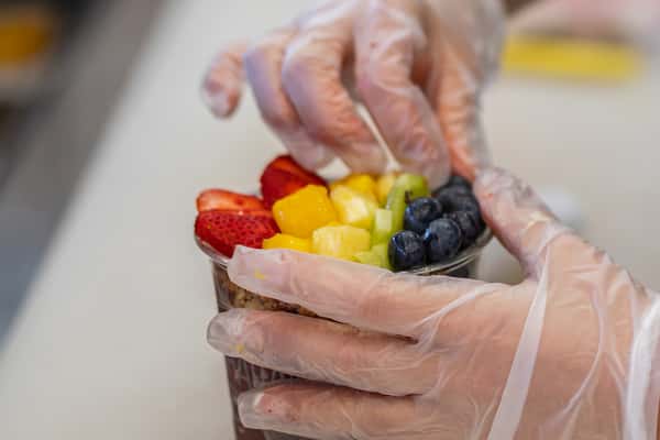 Gloved hands carefully arranging assorted fruits—strawberries, mango, pineapple, kiwi, and blueberries—on top of an acai bowl.
