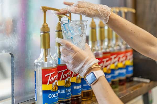 A gloved hand pumps blue raspberry syrup into an ice-filled cup from a row of Torani syrup dispensers.