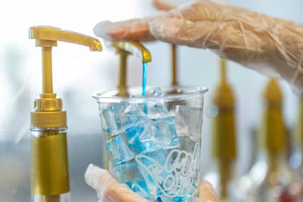 Blue syrup being pumped into a clear ALOHANA cup filled with ice by a gloved hand.