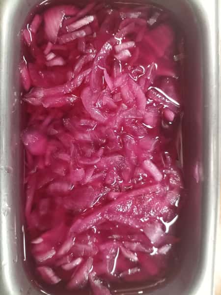 Container of vibrant pink pickled red onions soaking in brine, a popular tangy garnish for tacos, sandwiches, and salads.