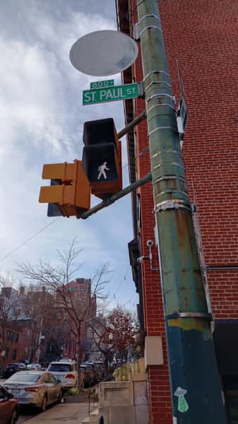 Urban street corner with pedestrian crossing signal and green 'St. Paul St' sign on a pole beside a red brick building in a city neighborhood.