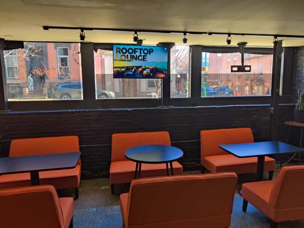 Modern casual restaurant lounge with orange chairs, small café tables, black brick walls, and large street-facing windows, with a TV display promoting a rooftop lounge.