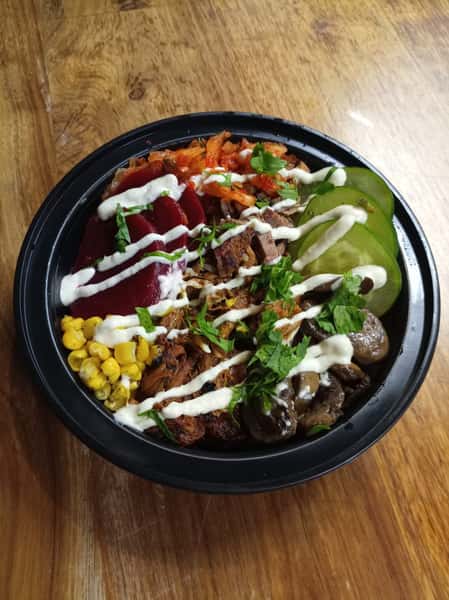Loaded BBQ grain bowl with smoked pulled pork, roasted corn, mushrooms, cucumbers, pickled vegetables, fresh herbs, and creamy sauce drizzle on a wooden table.