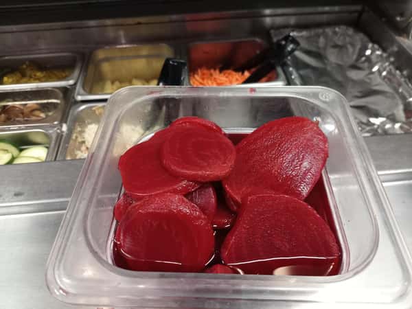 Clear food prep container filled with sliced pickled beets in brine on a restaurant prep counter with fresh ingredients in the background.