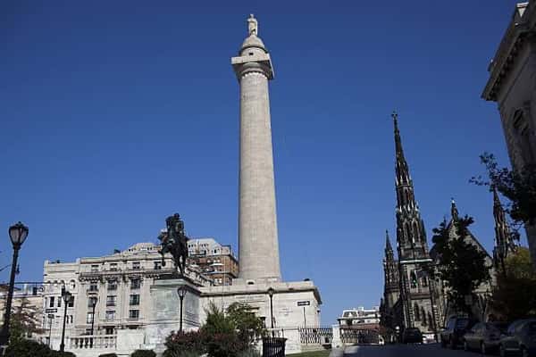 Washington Monument at Mount Vernon Place in Baltimore, featuring the tall historic column monument, surrounding statues, and nearby church spires against a clear blue sky.