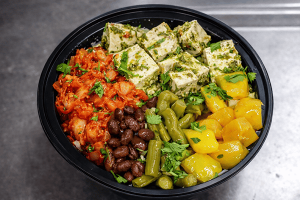 Colorful vegetarian grain bowl with herb-marinated tofu, kimchi, black beans, pickled vegetables, and fresh herbs over rice in a modern fast-casual restaurant bowl.