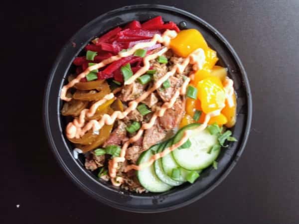 BBQ grain bowl with smoked pulled pork, pickled red beets, mango chunks, cucumber slices, jalapeños, and scallions over rice, finished with a spicy creamy sauce drizzle.