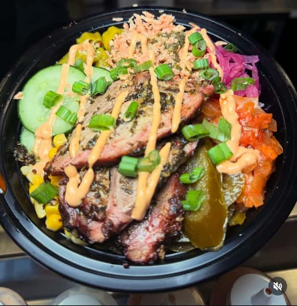 BBQ steak grain bowl topped with sliced smoked steak, pickled vegetables, cucumber, roasted corn, kimchi, and scallions, finished with a spicy creamy sauce drizzle in a fast-casual restaurant bowl.