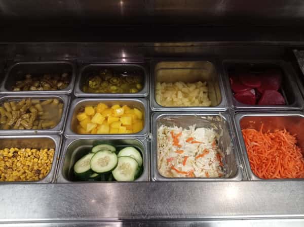 Restaurant prep station with stainless steel ingredient bar featuring toppings like roasted corn, cucumber slices, diced pineapple, jalapeños, pickled vegetables, shredded carrots, and coleslaw for customizable bowls and salads.
