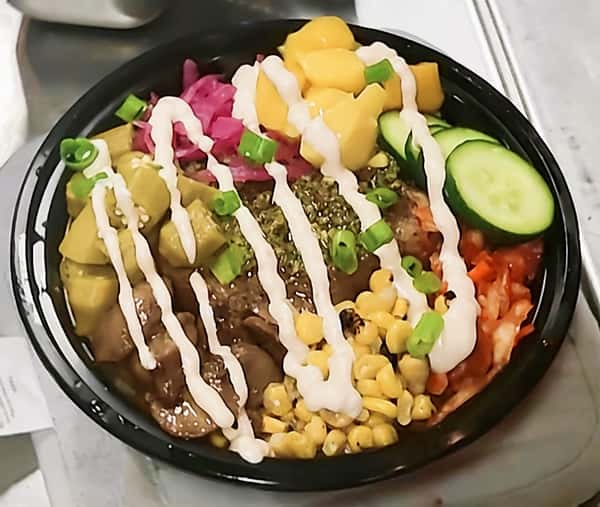 Grain bowl with grilled steak, avocado chunks, pickled red onions, mango salsa, roasted corn, cucumber slices, and scallions, drizzled with creamy sauce in a colorful modern restaurant bowl.