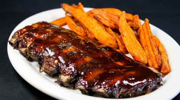 Rack of ribs covered in dark brown BBQ sauce, served with orange sweet potato fries on a white plate.