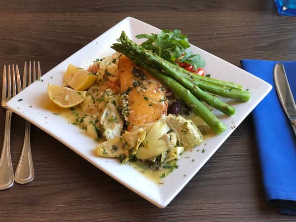 Grilled salmon with asparagus, orzo, artichokes, olives, and lemon wedges on a square white plate