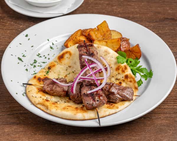 Beef souvlaki skewers over grilled pita topped with red onion, served with roasted potatoes and parsley