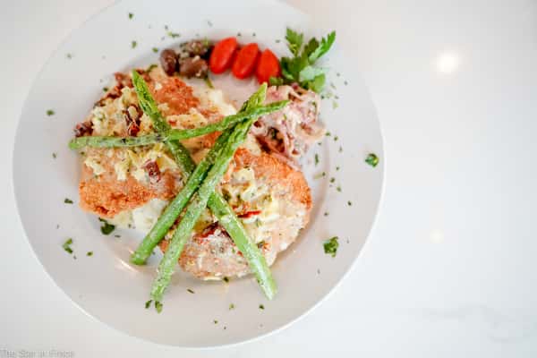 A plated seafood entrée featuring breaded salmon topped with a creamy sauce, served over mashed potatoes and crossed with asparagus spears, garnished with cherry tomatoes and herbs.