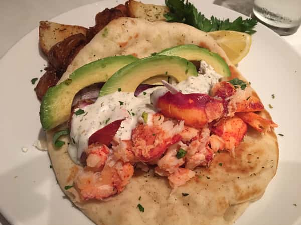 A lobster pita topped with chunks of lobster meat, sliced avocado, red onions, and creamy herb sauce, served with roasted potatoes and a lemon wedge on the side.