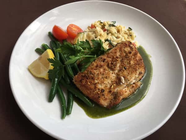 Pan-seared fish with green beans, orzo, cherry tomatoes, lemon wedge, and herb sauce on a white plate