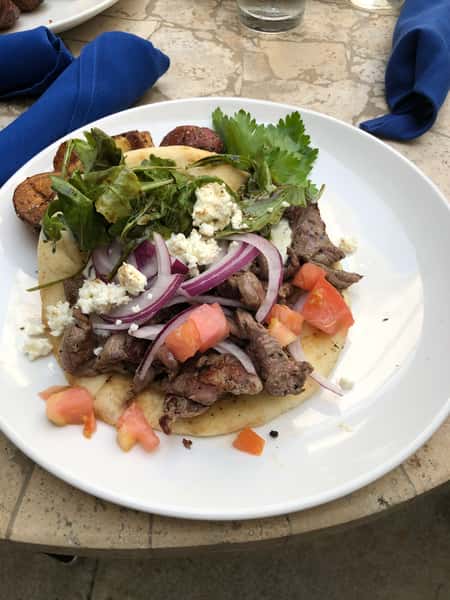 Gyro-style pita topped with grilled meat, feta, red onion, tomatoes, and mixed greens on a white plate