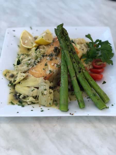 Pan-seared salmon topped with capers, served over artichokes in a light butter sauce, accompanied by grilled asparagus, cherry tomatoes, fresh parsley, and lemon wedges on a white square plate