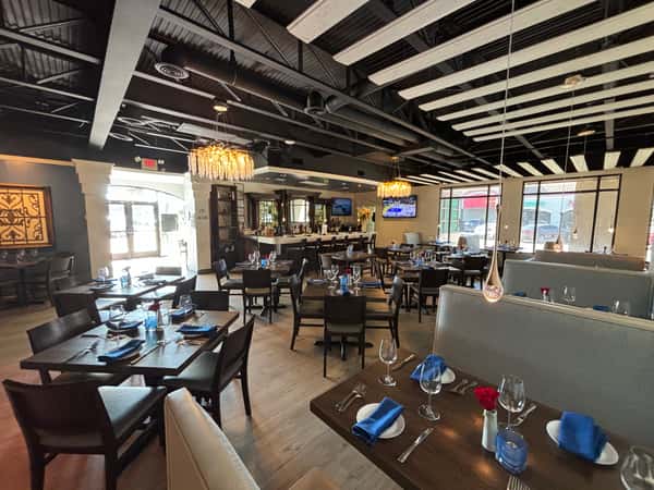 Modern restaurant dining room with wooden tables, blue napkins, chandeliers, and bar area in background