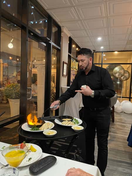 Server flambéing a dish tableside on a cart, flame rising from a pan, with pita and lemon wedges in a restaurant dining room