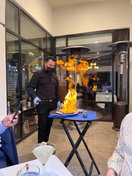 A server dressed in black prepares a flambé dish tableside, with tall flames rising from a pan on a serving tray. The scene is set inside a restaurant with glass walls, patio heaters, and diners seated in the background. A martini glass is visible on the table in the foreground as guests watch the presentation.