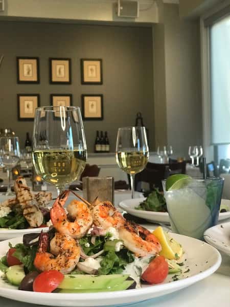 Grilled shrimp salad topped with feta, avocado, cherry tomatoes, and red onion, served with white wine and a lime cocktail in an elegant restaurant setting