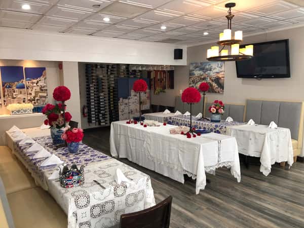 Private dining room with white tablecloths, red floral centerpieces, Greek artwork, and wine rack display