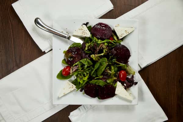 An overhead view of a beet salad arranged on a square white plate, featuring mixed greens, cherry tomatoes, triangular slices of feta, and round slices of roasted beets, drizzled with dressing and served with a knife on the side.