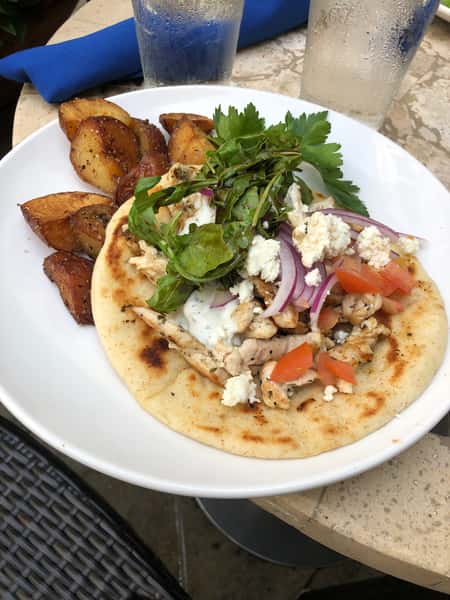 A grilled chicken pita topped with red onions, tomatoes, feta, mixed greens, and tzatziki sauce, served with roasted potato wedges on a white plate.