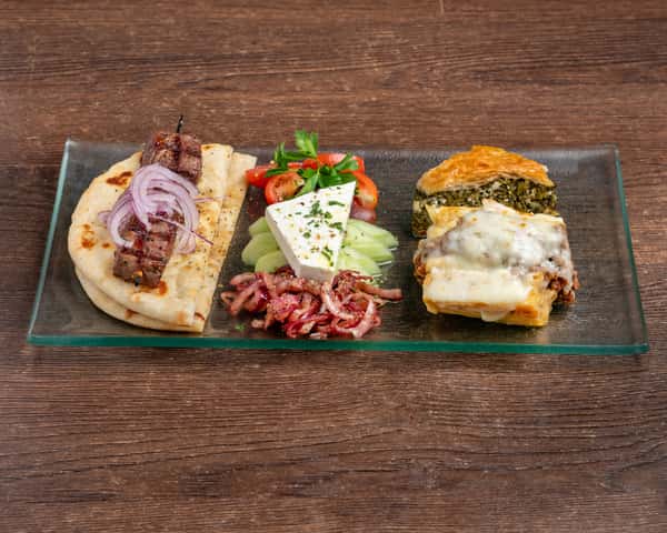 A Mediterranean-style sampler plate featuring a grilled beef skewer wrapped in pita with sliced red onions, a wedge of feta cheese with cucumbers and cherry tomatoes, marinated red onions, a slice of spinach pie (spanakopita), and a portion of moussaka topped with melted béchamel, all served on a rectangular glass plate.