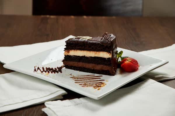 A slice of layered chocolate cake with dark and white cream fillings, topped with chocolate ganache and decorative drizzle, served on a square white plate with chocolate sauce accents and a fresh strawberry on the side.