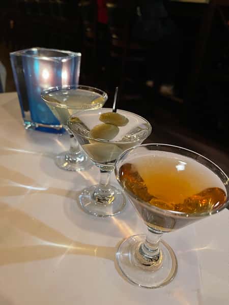 Three martinis lined up on a white tablecloth—one amber-colored, one classic with olives, and one pale—set beside a glowing blue candle holder in a dimly lit restaurant.