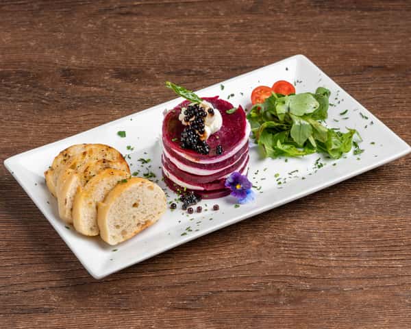 Plated beet and goat cheese stack layered in rounds, topped with crème and caviar, served with sliced baguette and a small side salad on a rectangular white plate
