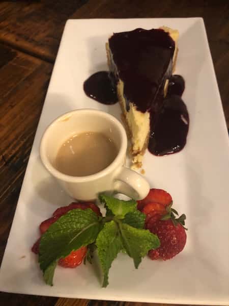 Slice of cheesecake topped with berry sauce, served with espresso and fresh strawberries on a white rectangular plate