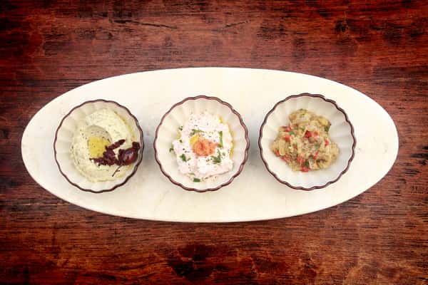 Three small bowls of Mediterranean dips including hummus, taramasalata, and eggplant spread on an oval platter