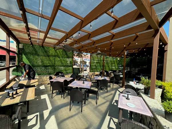 An outdoor patio dining area with multiple tables and woven chairs arranged under a wooden pergola. String lights hang overhead, and green hedge walls surround the space, creating a cozy atmosphere. The patio is decorated with potted plants and receives bright natural sunlight.