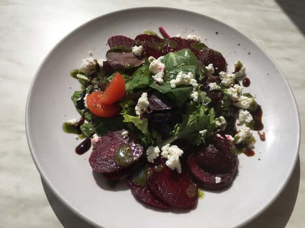 A beet salad topped with mixed greens, crumbled feta, cherry tomatoes, and drizzled with balsamic glaze and herb dressing on a white plate.