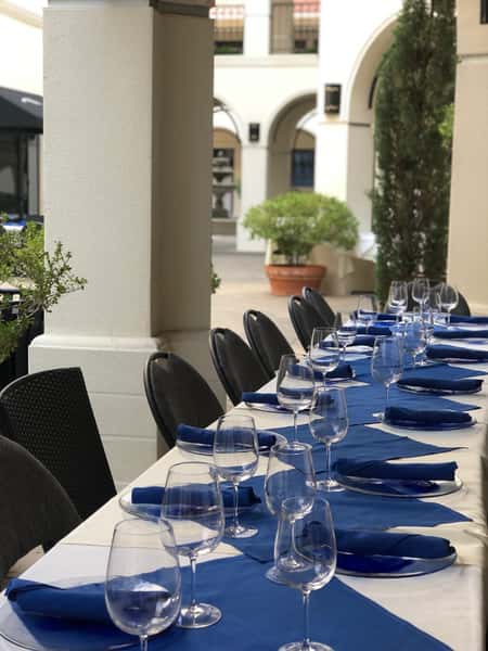 Outdoor patio table set with blue runners, folded napkins, wine glasses, and black chairs near arched courtyard