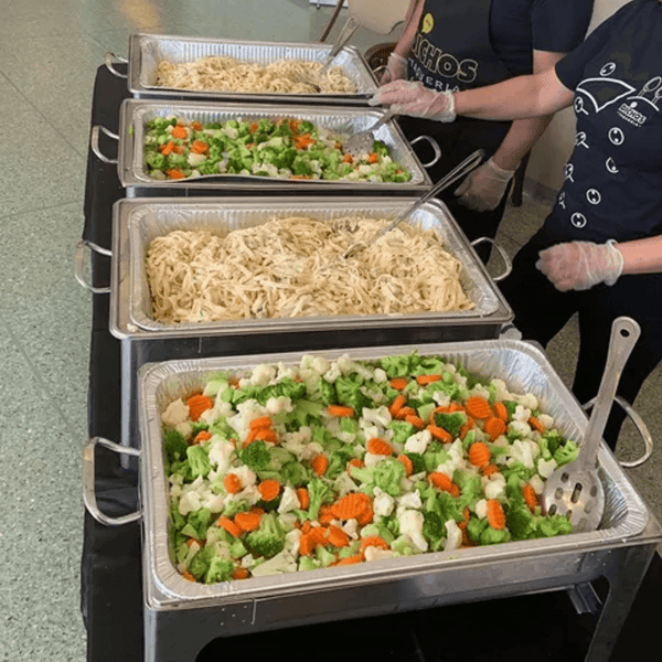Catering setup from Dichos Taqueria featuring large trays of shredded chicken and steamed mixed vegetables including broccoli, carrots, and cauliflower, served buffet-style by staff wearing branded uniforms and gloves.
