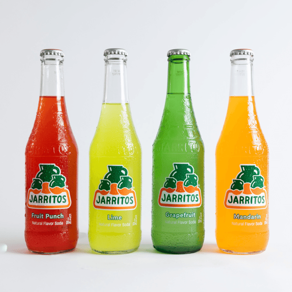 Four colorful glass bottles of Jarritos soda in fruit punch, lime, grapefruit, and mandarin flavors, arranged side by side on a white background, showcasing popular Mexican soft drinks.