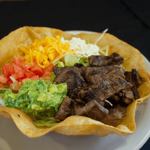 Taco salad served in a crispy tortilla bowl, filled with grilled steak, shredded lettuce, guacamole, diced tomatoes, shredded cheese, and sour cream — a hearty Mexican-style entrée.