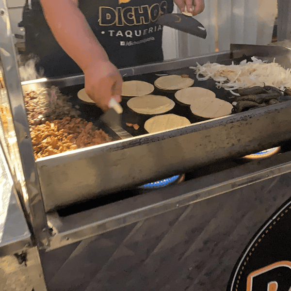 Street food vendor from Dichos Taqueria grilling fresh corn tortillas, seasoned meat, onions, and jalapeños on a flat top grill — authentic Mexican taco preparation in progress.