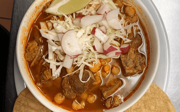 Traditional Mexican red pozole served in a white bowl, filled with hominy, tender pork chunks, and garnished with shredded cabbage, sliced radishes, and a wedge of lime, accompanied by crispy tostadas.