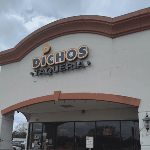 Exterior of Dichos Taqueria, showcasing the restaurant's bold logo with a speech bubble icon above the name, set on a stucco building with arched trim under a partly cloudy sky.