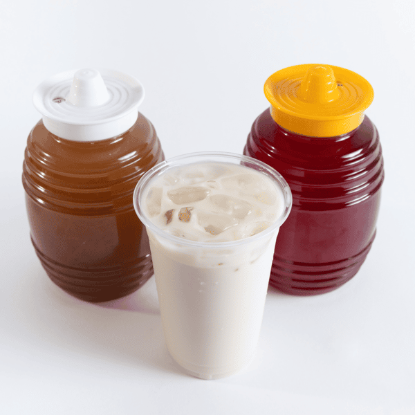 Three traditional Mexican aguas frescas, featuring a creamy horchata in a clear cup filled with ice, flanked by dispensers of tamarindo (brown) and jamaica (hibiscus, red) on a white background.