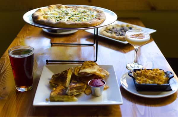 Pizza on serving tray, side of mac and cheese, grilled sandwiches, chips and beer
