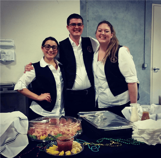 3 employees wearing white dress shirts and black vests, standing behind a catering display decorated with mardi gras decorations